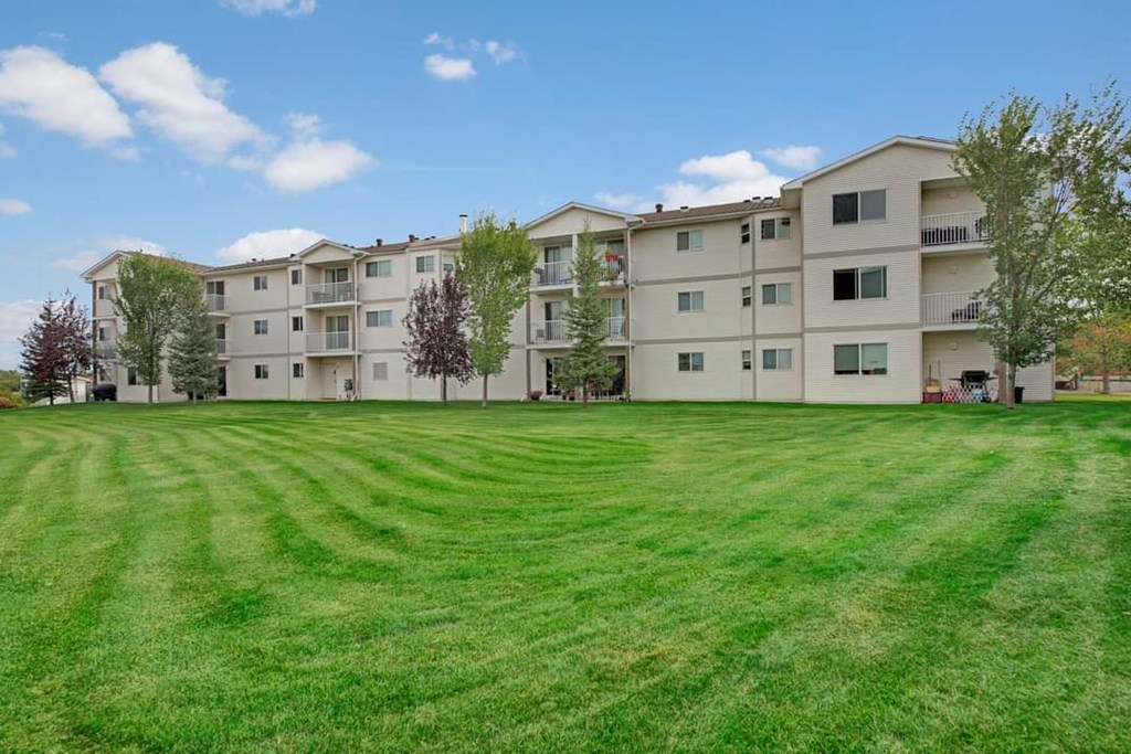 Four-story beige Aspen Terrace Apartment Homes building with balconies, surrounded by neatly mowed green lawn and trees. Bright, clear sky enhances a serene atmosphere.