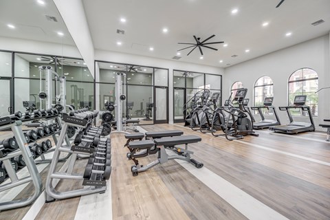 A modern fitness center here at Desert Club featuring wood-style flooring, mirrored walls, and bright overhead lighting. The space includes free weights, adjustable benches, stationary bikes, and cardio machines arranged for convenience. Large windows and glass-enclosed training areas create an open, motivating environment for daily workouts.