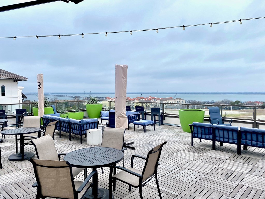 Rooftop patio here at Florence at the Harbor Apartment Homes with blue cushioned seating, green planters, and round tables under string lights. Overcast sky, distant view of buildings and water.