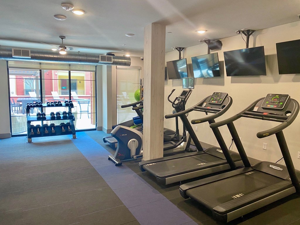 Modern gym here at Florence at the Harbor Apartment Homes with treadmills, exercise bike, free weights, and TVs on the wall. Large windows allow natural light, creating an inviting atmosphere.