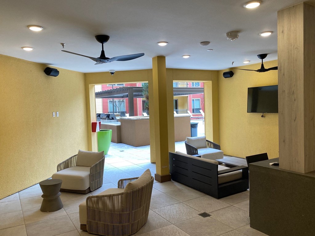 Modern lounge area here at Florence at the Harbor Apartment Homes with yellow walls, cozy seating, a wall-mounted TV, overhead ceiling fans, and a view of an outdoor patio with vibrant planters.