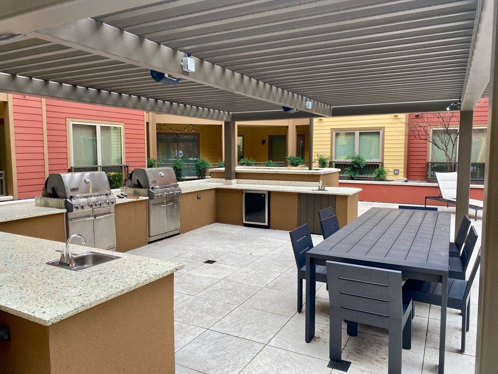Outdoor patio here at Florence at the Harbor Apartment Homes with modern grills under a pergola. Granite countertops feature a sink. A sleek black dining table with chairs is placed nearby.