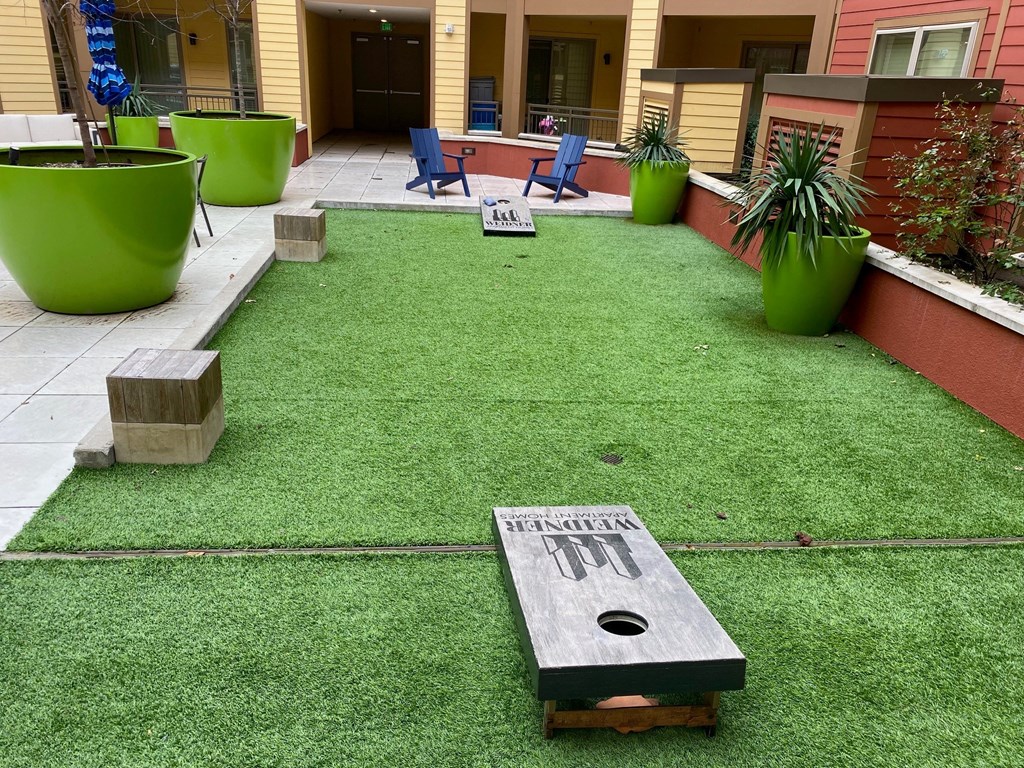 Artificial grass courtyard here at Florence at the Harbor Apartment Homes with cornhole boards, surrounded by large green planters and colorful chairs. The setting feels inviting and playful.
