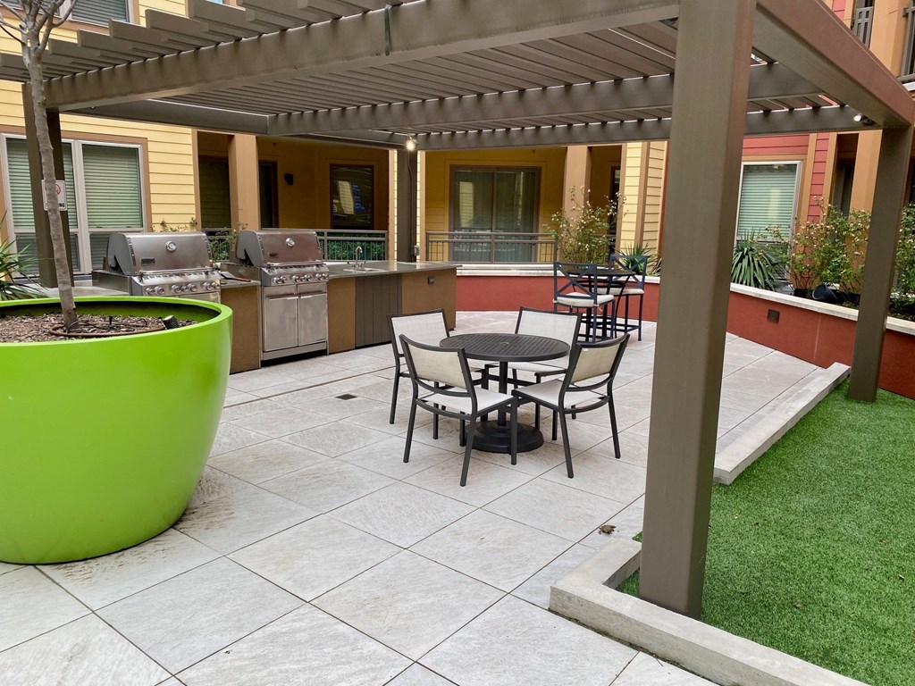 Outdoor patio area here at Florence at the Harbor Apartment Homes featuring a pergola, two stainless steel grills, patio tables with chairs, a large green planter, and surrounding plants. Cozy ambiance.