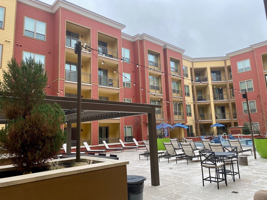 Modern courtyard here at Florence at the Harbor Apartment Homes with a pool, lounge chairs, and tables. Buildings are red and yellow with balconies. Overcast sky and string lights create a cozy ambiance.