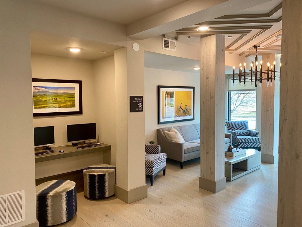 Modern lounge here at Florence at the Harbor Apartment Homes with a cozy ambiance. Two computer stations with stools on the left and a seating area on the right, featuring a sofa and chairs. A chandelier hangs from the ceiling, and artwork decorates the walls, adding a touch of elegance.