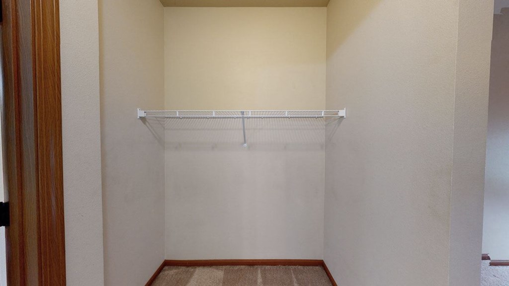 Empty beige closet here at Grand River Estates Apartment Homes with a wire shelf mounted on the back wall. The doorframe is wooden, and the floor is carpeted. Simple, tidy space.
