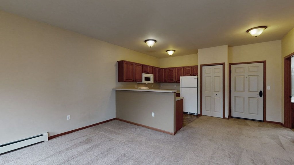 Interior of an empty apartment here at Grand River Estates Apartment Homes featuring a carpeted living area and a kitchen. The kitchen includes wooden cabinets, a white fridge, and a microwave. The walls are beige, and three ceiling lights brighten the space, creating a warm, inviting atmosphere.
