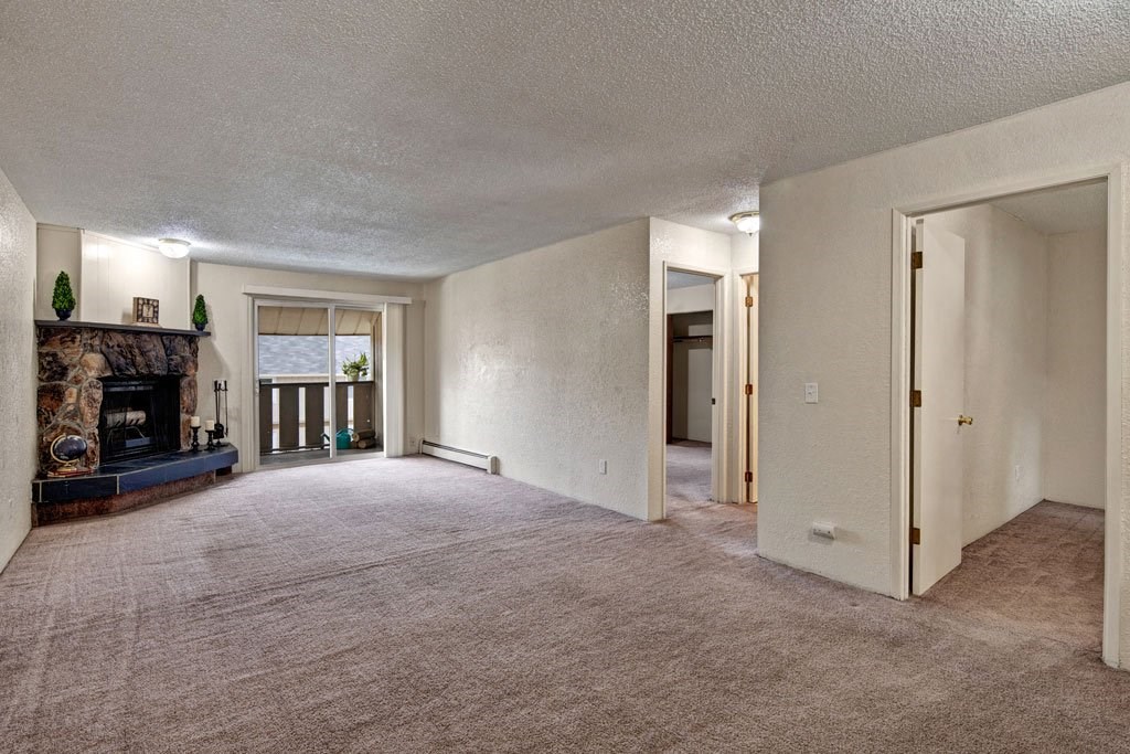 Spacious living room here at Greenbriar Apartments with light beige carpet, stone fireplace, sliding glass doors, and open doorways. Cozy and inviting with natural light.