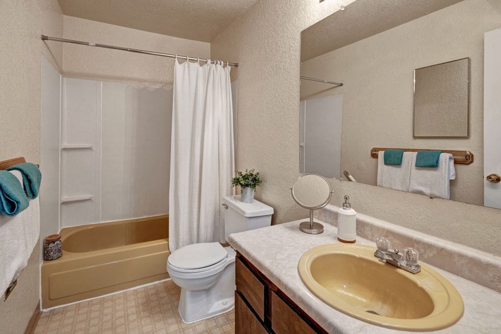 Bathroom here at Greenbriar Apartments with a yellow bathtub and sink, white shower curtain, and countertop. Towels and a small plant add color. Clean and neutral decor.