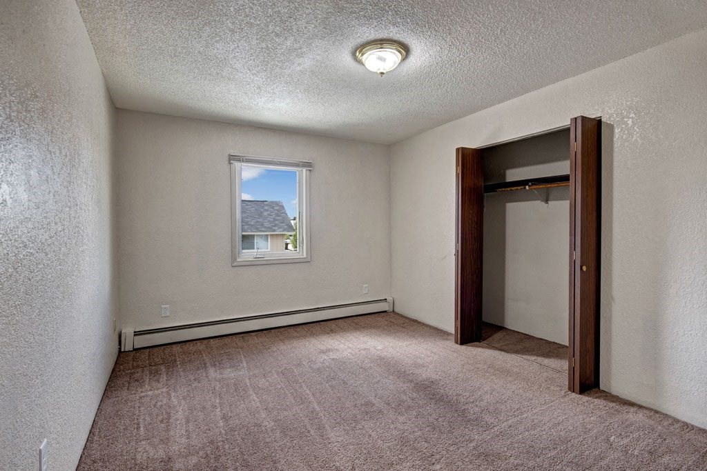 Bedroom here at Greenbriar Apartments with beige walls and brown carpet. A single window shows a roof and sky. Open closet with wooden doors on the right. Simple and neutral tone.