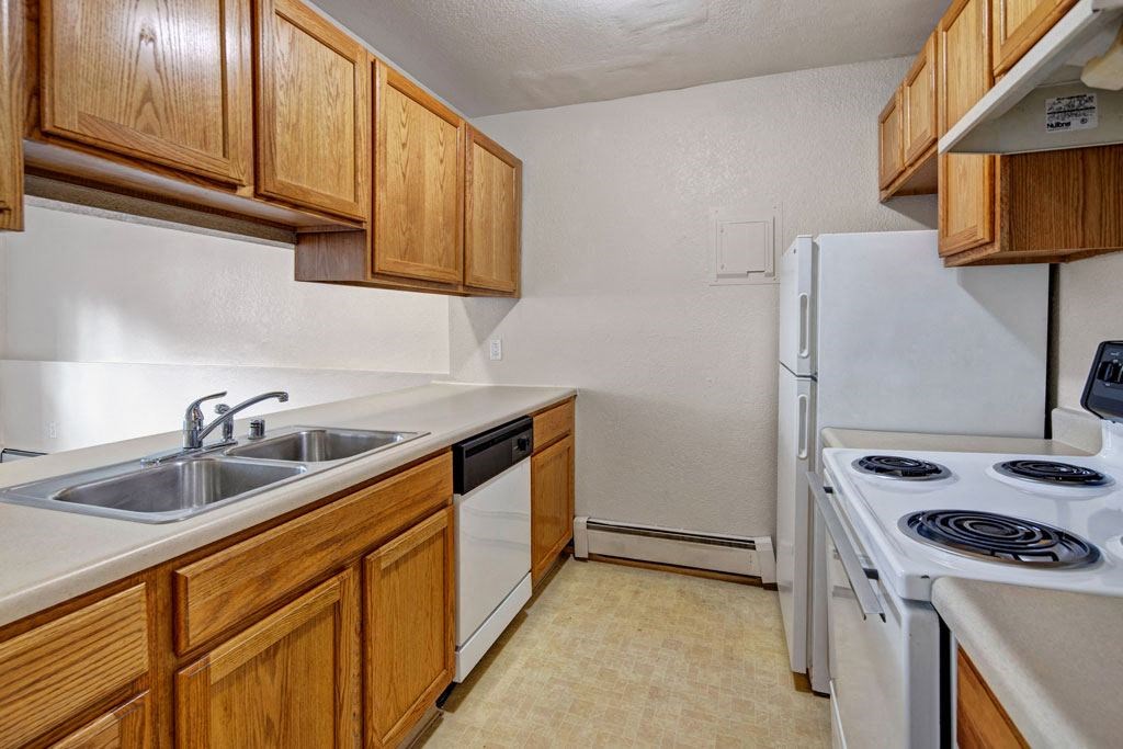 Narrow kitchen here at Greenbriar Apartments with wooden cabinets, beige countertops, and appliances including a stainless-steel sink, dishwasher, white stove, and fridge. Cozy and functional.