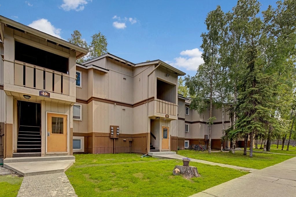 Two-story beige apartment buildings here at Greenbriar Apartments with brown trim amidst a landscaped, grassy area with trees and blue sky, conveying tranquility.