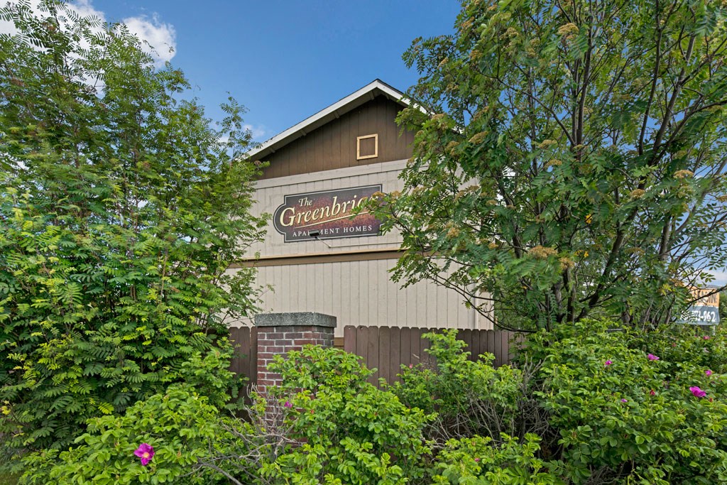 A building sign for "The Greenbriar Apartment Homes" is partially obscured by lush green trees and shrubs, with a clear blue sky in the background.