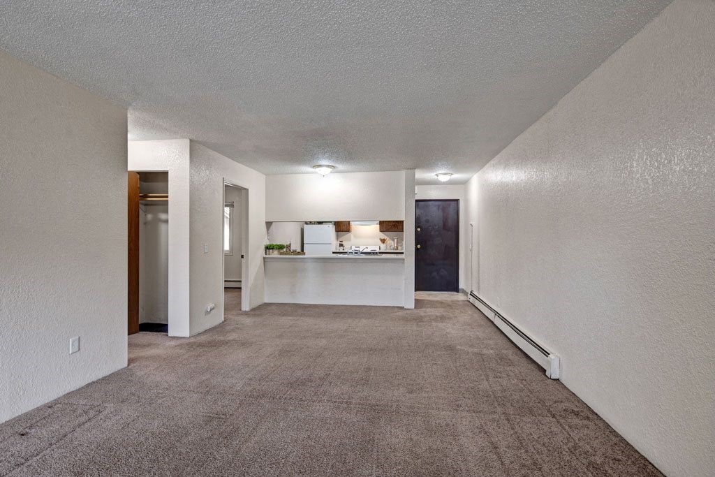 Empty living room here at Greenbriar Apartments with beige carpet and white walls, featuring a small closet on the left, a kitchen with a counter in the middle, and a dark wooden door on the right. Bright overhead lighting creates a clean, spacious feel.