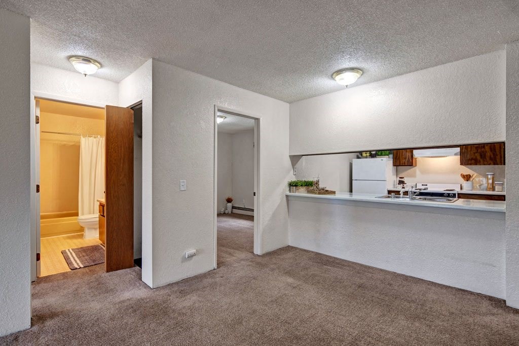 Cozy apartment interior here at Greenbriar Apartments with beige carpet and white walls. Features a small kitchen with a breakfast bar, bathroom with shower curtain, and softly lit rooms.