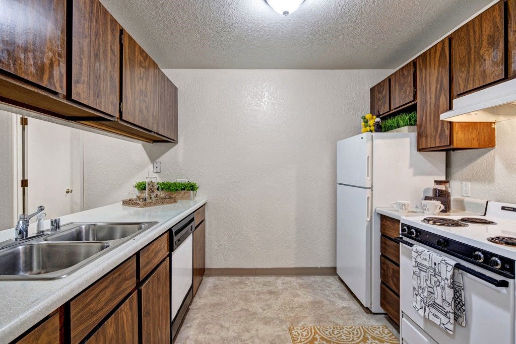Compact kitchen here at Greenbriar Apartments with dark wood cabinets, white appliances including a fridge and stove, dual sink, and decorative green plants. Cozy and functional.