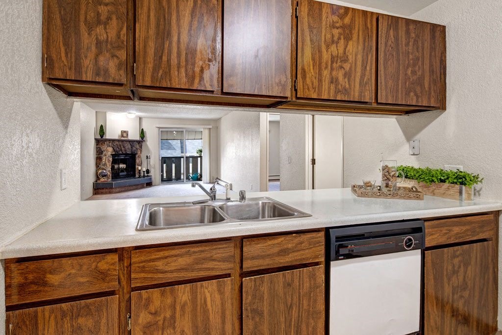 Stylish kitchen here at Greenbriar Apartments with wooden cabinets, double sink, and dishwasher. A pass-through window reveals a cozy living room with a fireplace, creating a warm tone.