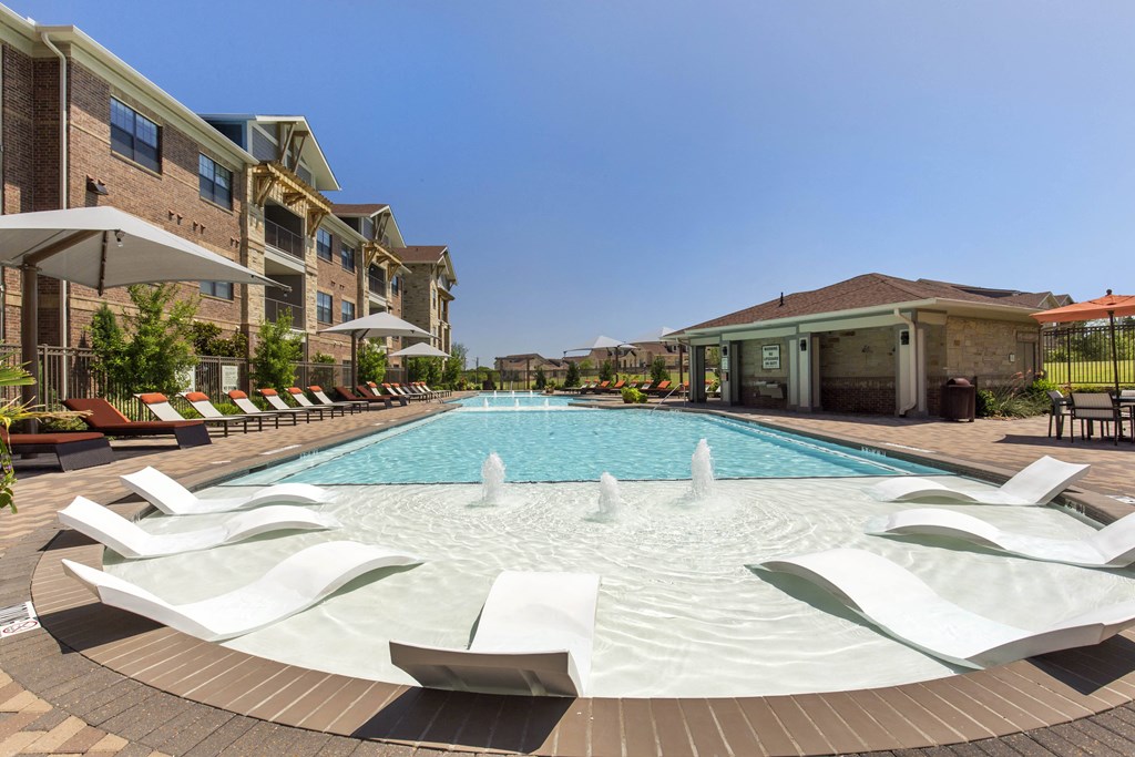 A luxurious pool area here at Sorrel Fairview Apartment Homes with clear blue water, surrounded by brick buildings and sun loungers. White deck chairs rest in shallow water, under a clear sky.