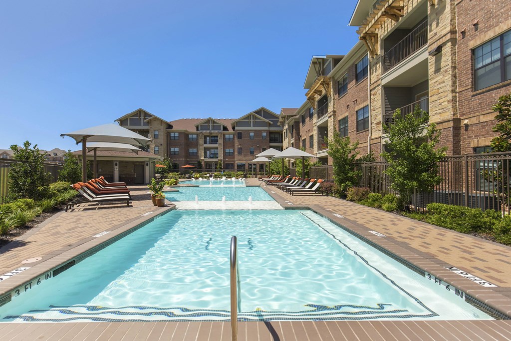 Outdoor pool area here at Sorrel Fairview Apartment Homes with clear water, surrounded by lounge chairs and umbrellas. Brick apartment buildings in the background under a clear blue sky.