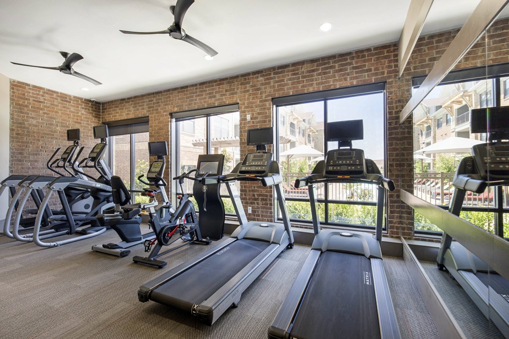A modern gym here at Sorrel Fairview Apartment Homes with brick walls, featuring treadmills, stationary bikes, and elliptical machines. Large windows offer a view of a sunny courtyard.