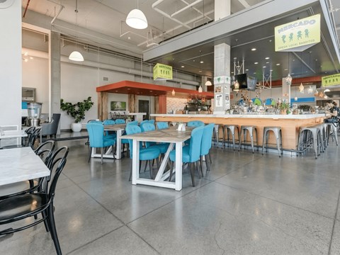 A modern communal dining and bar area here at Blue and Lime featuring polished concrete floors, a long wood-accented bar with metal stools, and a central dining table surrounded by bright blue upholstered chairs. Industrial lighting, exposed ceilings, and greenery create an open, social atmosphere designed for gatherings and casual dining.