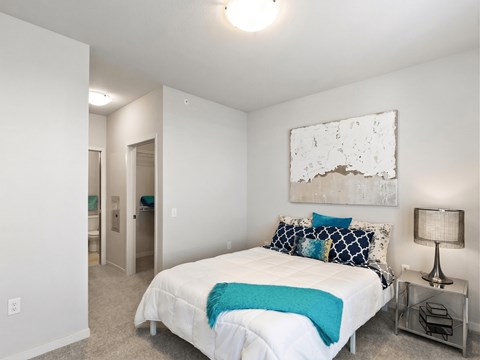 A serene bedroom here at Blue and Lime featuring a comfortable bed dressed in white bedding with blue and teal accent pillows and a matching throw. Neutral walls, soft carpeting, and a modern bedside table with a lamp create a calm atmosphere, while the open layout connects to a nearby closet and bathroom area for everyday convenience.