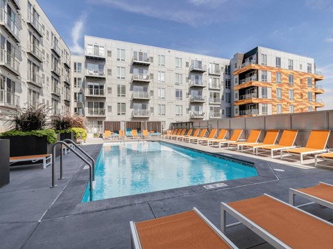 A resort-style outdoor pool here at Blue and Lime featuring clear water surrounded by a spacious deck lined with orange lounge chairs and modern planters. Contemporary apartment buildings frame the courtyard, while railings, steps, and open sky create a bright, inviting setting designed for relaxation and social gathering.