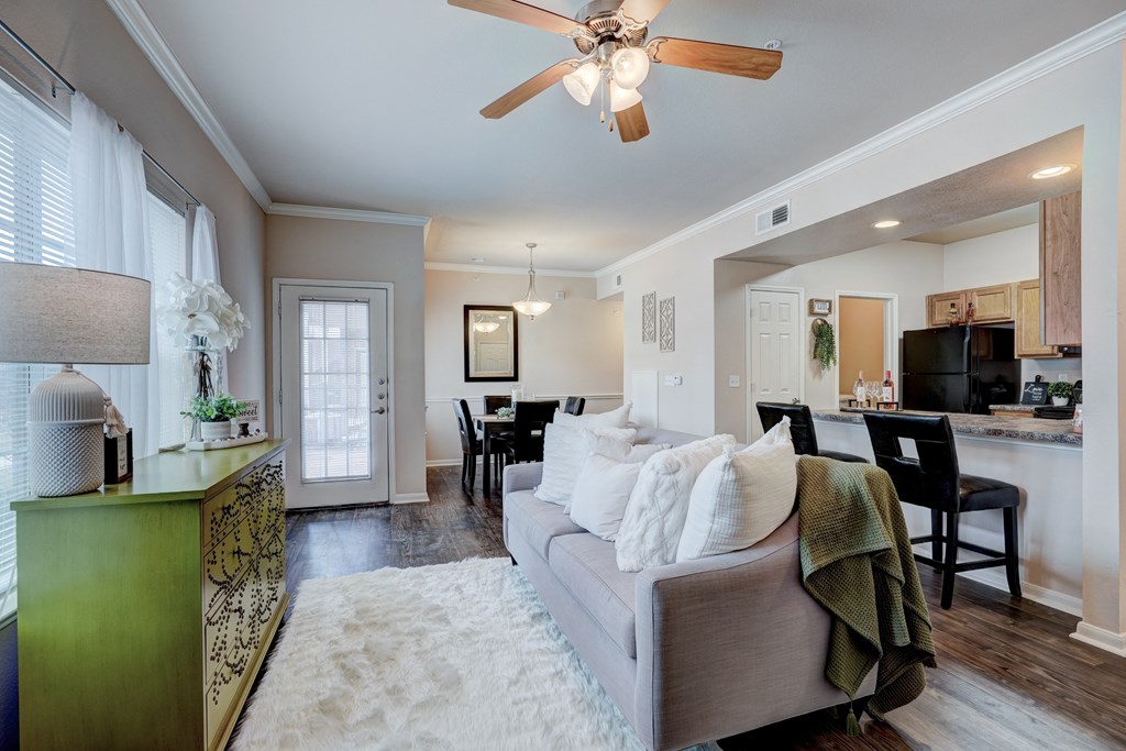 Cozy living room here at Lincoln at Central Park Apartment Homes with a ceiling fan, neutral-colored sofa, fluffy rug, and bright window. Green console table adds color. Dining area visible in the background.