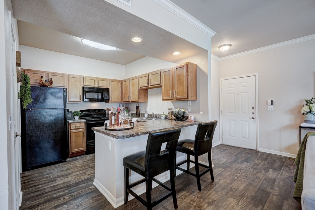 Cozy kitchen here at Lincoln at Central Park Apartment Homes with wooden cabinets and dark appliances. A counter with barstools faces the entrance. Warm lighting creates an inviting ambiance.