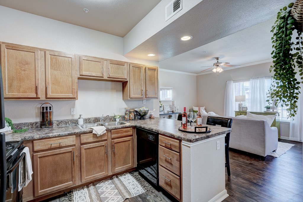 A modern kitchen here at Lincoln at Central Park Apartment Homes with wooden cabinets and marble countertops opens into a cozy living room. Natural light, plants, and soft decor create a welcoming atmosphere.
