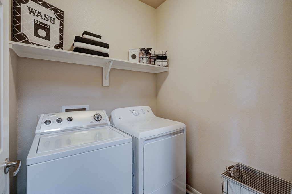 Compact laundry room here at Lincoln at Central Park Apartment Homes with beige walls featuring a white washer and dryer side by side. A shelf above holds folded towels, a sign, and cleaning supplies.