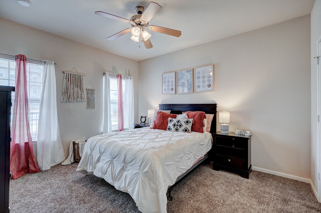 Cozy bedroom here at Lincoln at Central Park Apartment Homes with a ceiling fan, a neatly made bed with white and red pillows, two bedside tables with lamps, and soft lighting. Neutral tones create a calm ambiance.