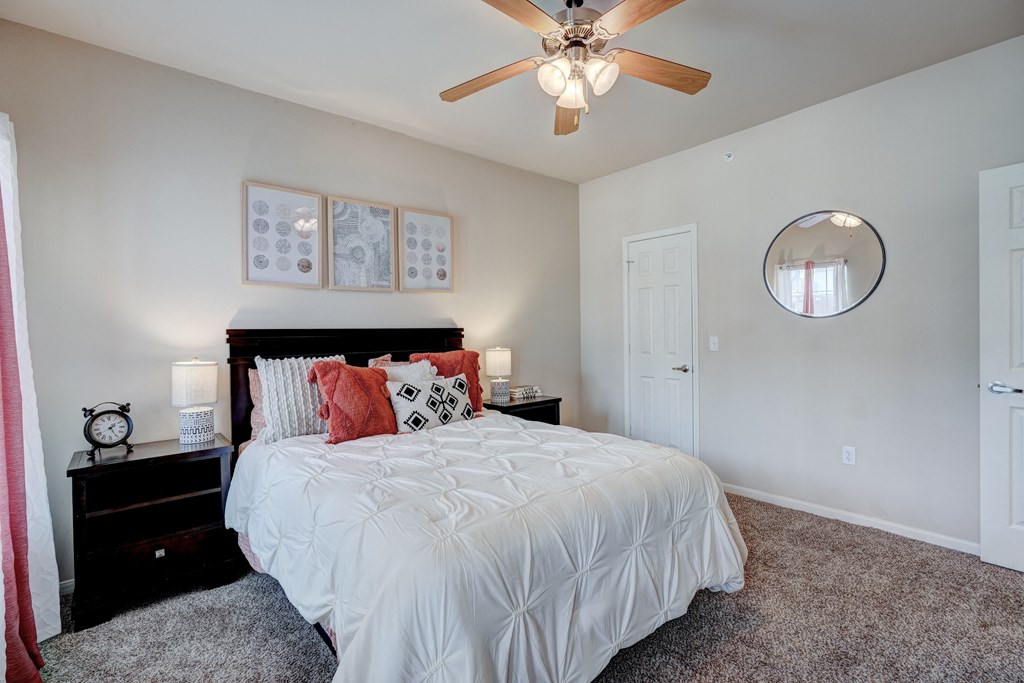 A cozy bedroom here at Lincoln at Central Park Apartment Homes with a white comforter, decorative pillows, and bedside tables. A ceiling fan overhead and a round mirror on a beige wall add warmth.