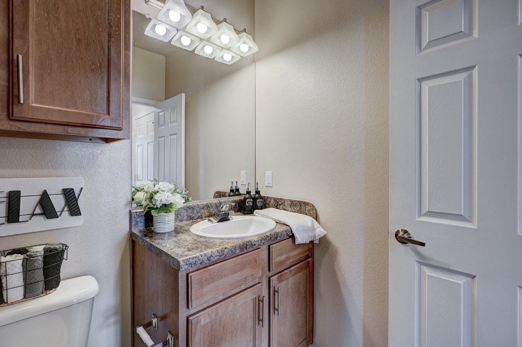Cozy bathroom here at Lincoln at Central Park Apartment Homes with a wood cabinet, granite countertop, white sink, flowers, and a towel. A "RELAX" sign and soft lighting convey a tranquil vibe.