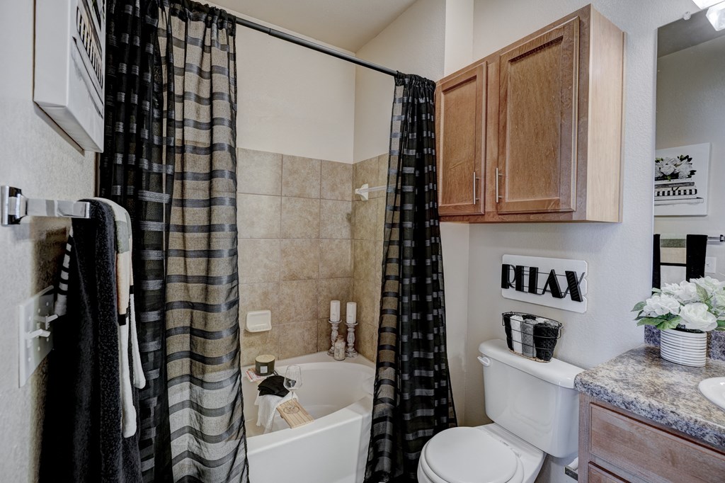 Compact bathroom here at Lincoln at Central Park Apartment Homes with dark plaid shower curtain, tiled bathtub, wooden cabinets, and "RELAX" sign above toilet. Tone is calm and inviting.