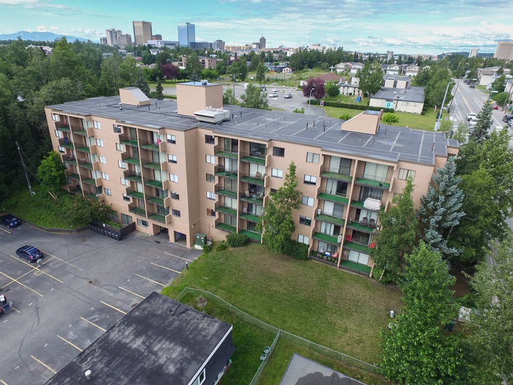 The Outlook Apartment - Exterior Aerial view of a beige, five-story Outlook apartment building with balconies, surrounded by green trees and a parking lot. City skyline in the distance under a cloudy blue sky.