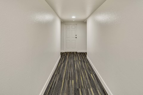 Narrow, brightly lit hallway at La Maisonnette Apartments with white walls and door at the end. The floor features dark patterned carpeting with subtle yellow stripes, creating a modern feel.