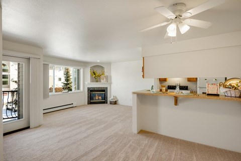 Bright living room here at La Maisonnette Apartments with beige carpet, a fireplace, large windows, and a kitchen bar. Soft, neutral tones create a cozy, welcoming atmosphere.
