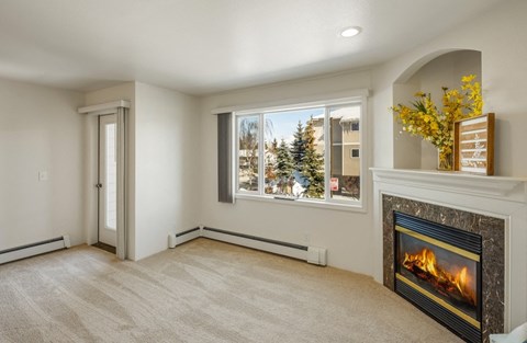 Bright living room here at La Maisonnette Apartments with plush carpet, large window showing snowy outside, and a modern fireplace. Yellow flowers and decor on mantel creating a cozy ambiance.