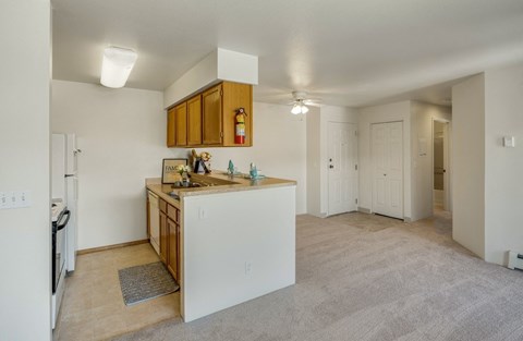 Cozy apartment interior here at La Maisonette Apartments featuring a small kitchen with wooden cabinets, beige countertops, and a dining bar. Adjacent is a carpeted living space, with closed white-paneled doors and visible lighting, creating a warm and inviting atmosphere.