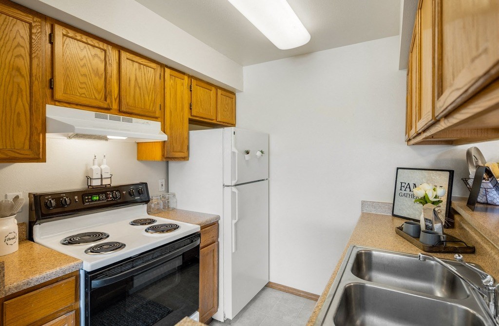 Compact kitchen here at La Maisonnette Apartments with wooden cabinets, a white stove, and fridge. Stainless steel sink in the foreground, decor includes a “Family” sign and flowers.