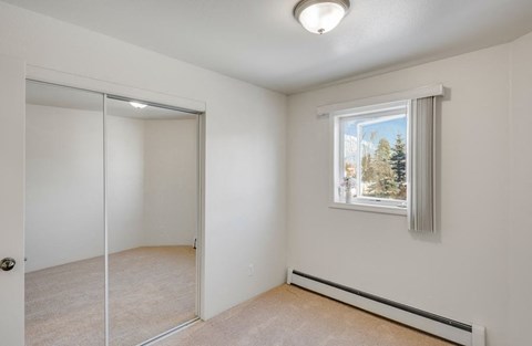 Small bedroom here at La Maisonnette Apartments with beige carpet and white walls, featuring a window with vertical blinds, a ceiling light, and a mirrored closet door reflecting light.