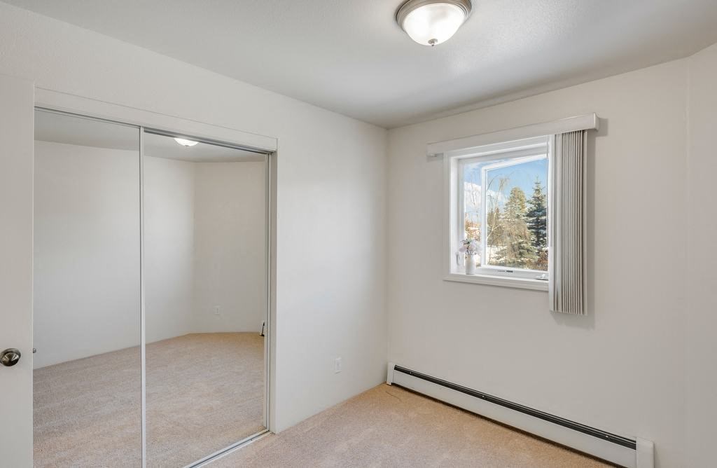 La Maisonnette Apartments - Bedroom Small bedroom here at La Maisonnette Apartments with beige carpet and white walls, featuring a window with vertical blinds, a ceiling light, and a mirrored closet door reflecting light.