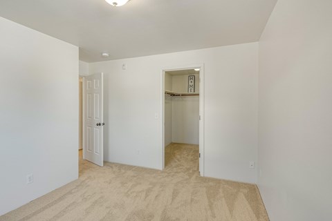 Bright bedroom here at La Maisonnette Apartments with beige carpet and white walls. Features an open closet with a "HOME" sign, and an ajar door leading to another room. Calm atmosphere.