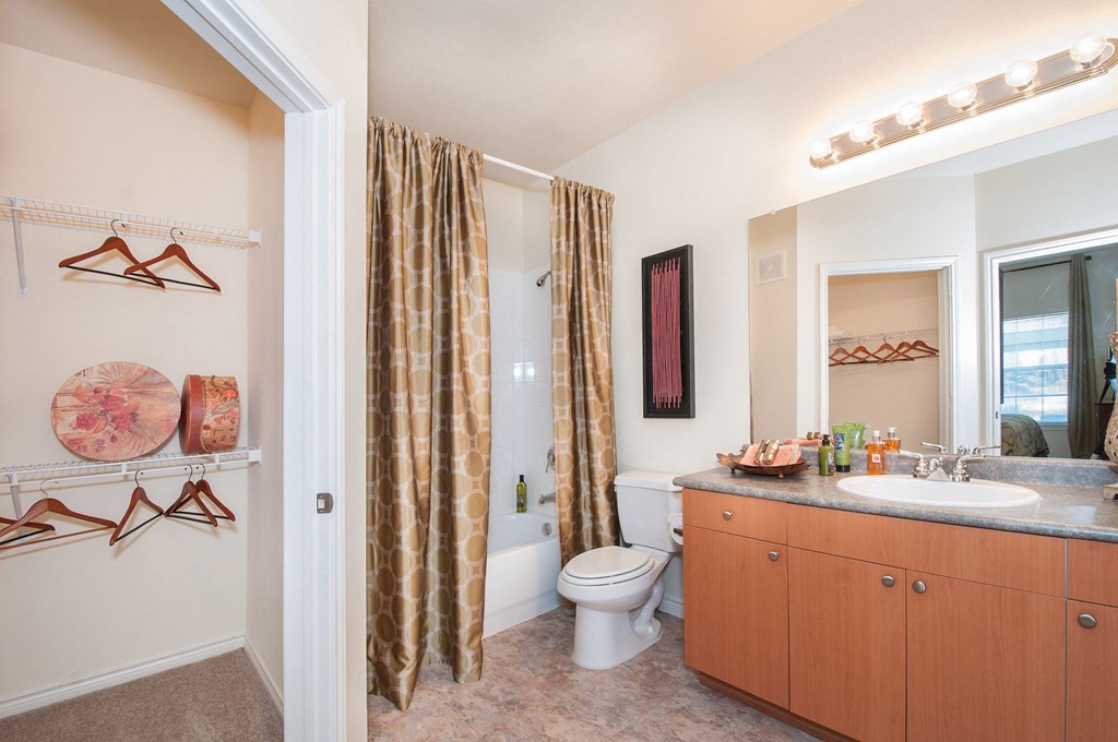Bathroom here at Landing at Mansfield Apartment Homes with wooden cabinets, beige patterned shower curtain, and bright vanity lights. Open closet with wooden hangers and decorative boxes.