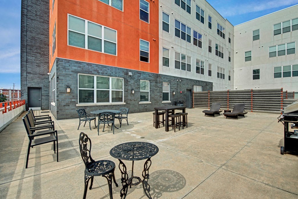 Patio at the Latitude Apartments with metal tables and chairs, loungers, and a barbecue grill. Surrounding buildings feature orange and grey facades under a clear sky.