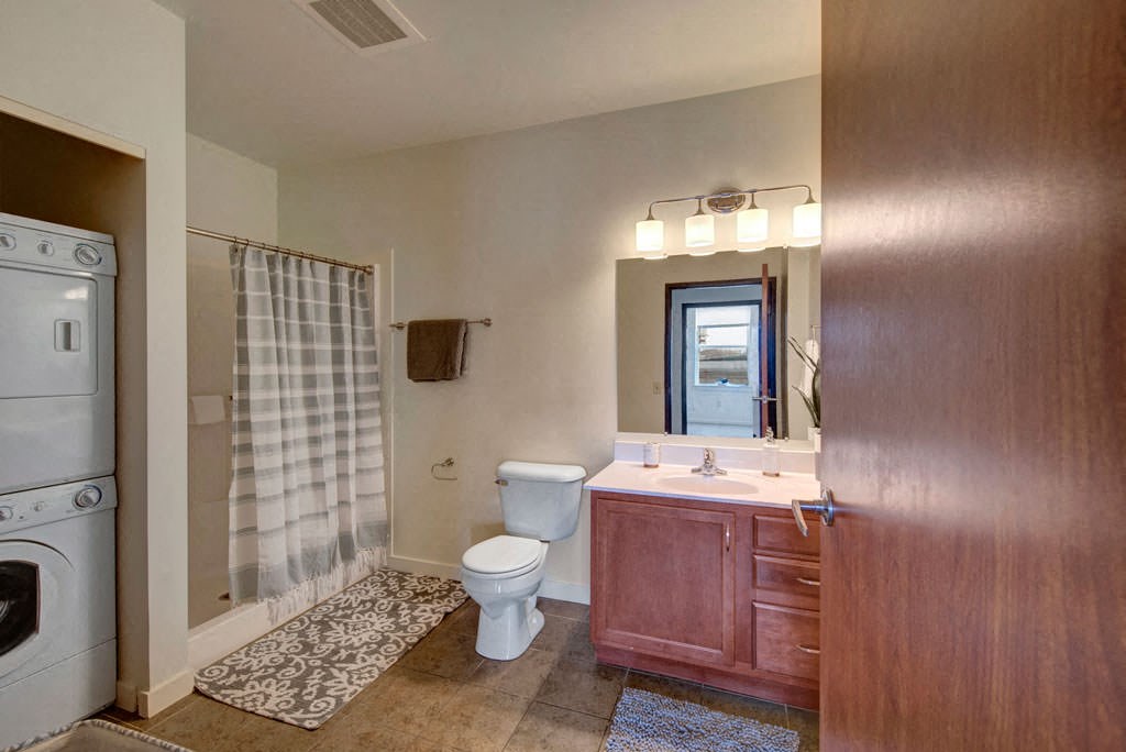 Modern bathroom at the Latitude Apartments with light wood cabinetry, a large mirror, and warm lighting. A striped shower curtain is adjacent to a stacked washer and dryer.