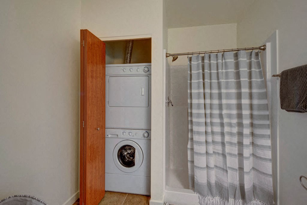 The Latitude Apartments bathroom with a striped shower curtain concealing a shower area. A stackable washer and dryer are inside an open wooden closet.