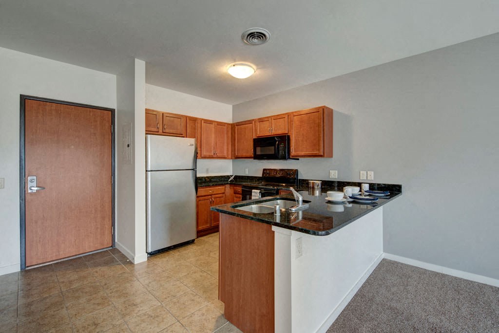 Modern kitchen at Latitude Apartments with stainless steel fridge, wooden cabinets, and a black granite countertop. Includes a built-in sink, microwave, and oven. Warm, inviting tone.